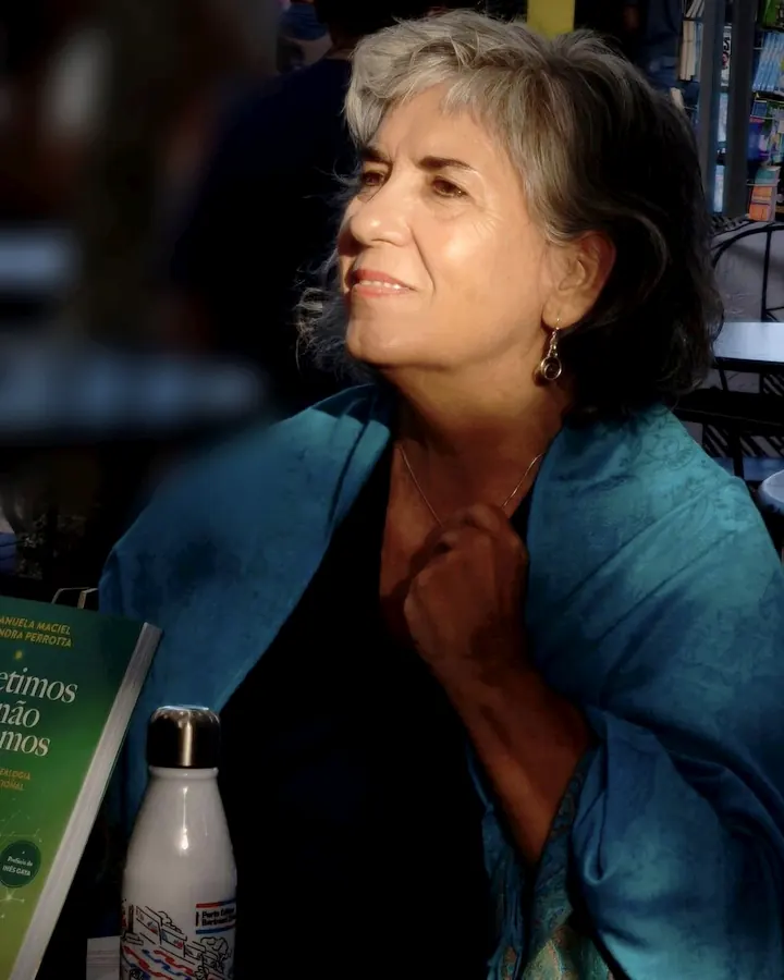 Manuela Maciel sentada ao ar livre, com um xaile azul, ao lado de um exemplar do livro Repetimos o que não reparamos.