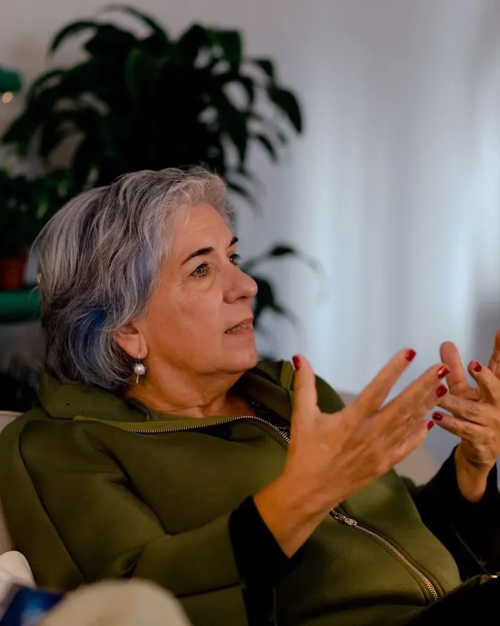 Manuela Maciel a conversar, sentada num sofá claro, com as mãos abertas em gesto e plantas ao fundo.